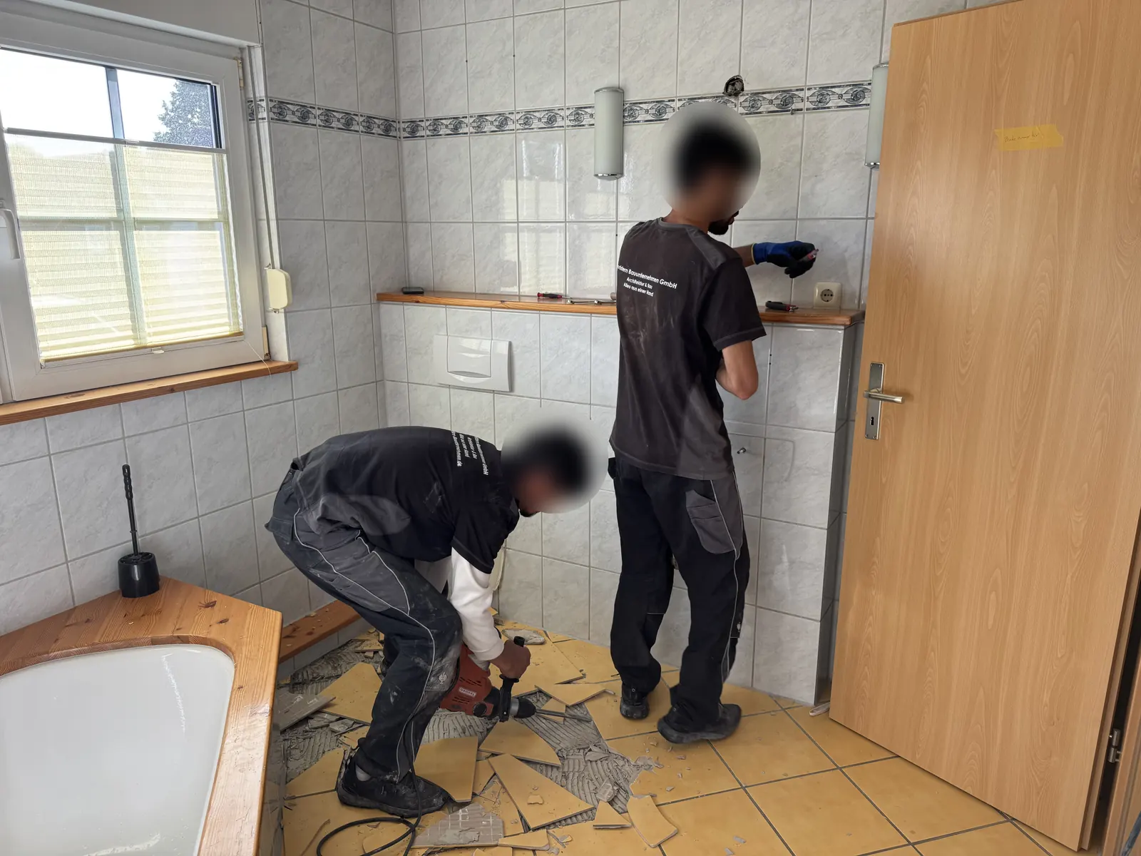 Zwei Handwerker entfernen in einem Badezimmer die alten Wand- und Bodenfliesen, auf dem Boden liegen abgeschlagene Fliesen