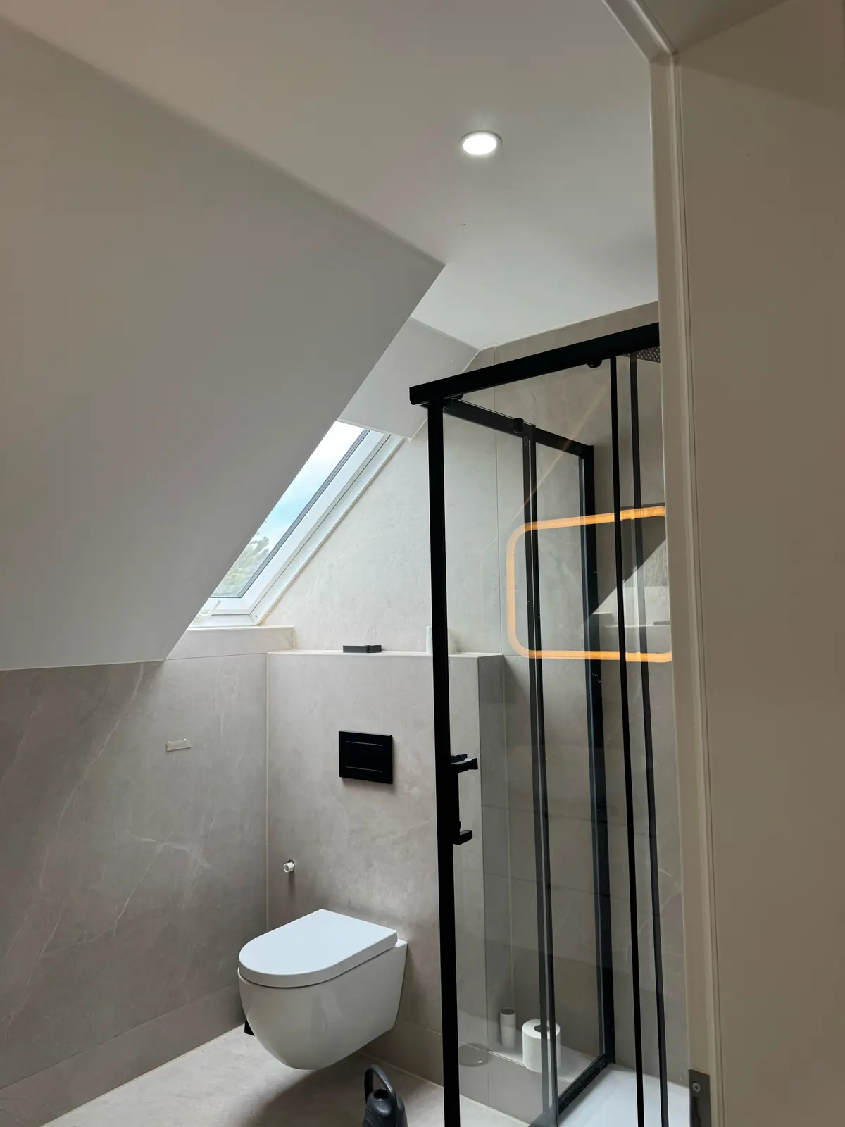 Fertiges Bad unter einer Dachschräge in Hamburg-Poppenbüttel mit Dachfenster, großformatigen hellgrauen Fliesen, Wand-WC mit schwarzer Betätigungsplatte und Duschkabine mit schwarzem Rahmen