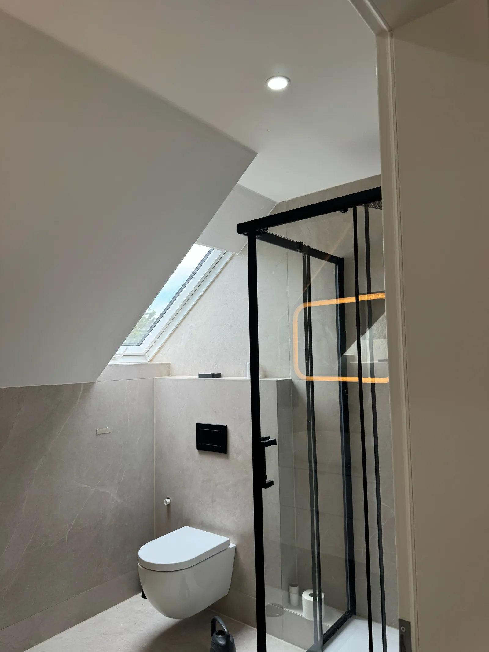 Fertiges Bad unter der Dachschräge mit Dusche, schwarz gerahmter Duschkabine, Wand-WC, Dachfenster und großformatigen hellgrauen Fliesen