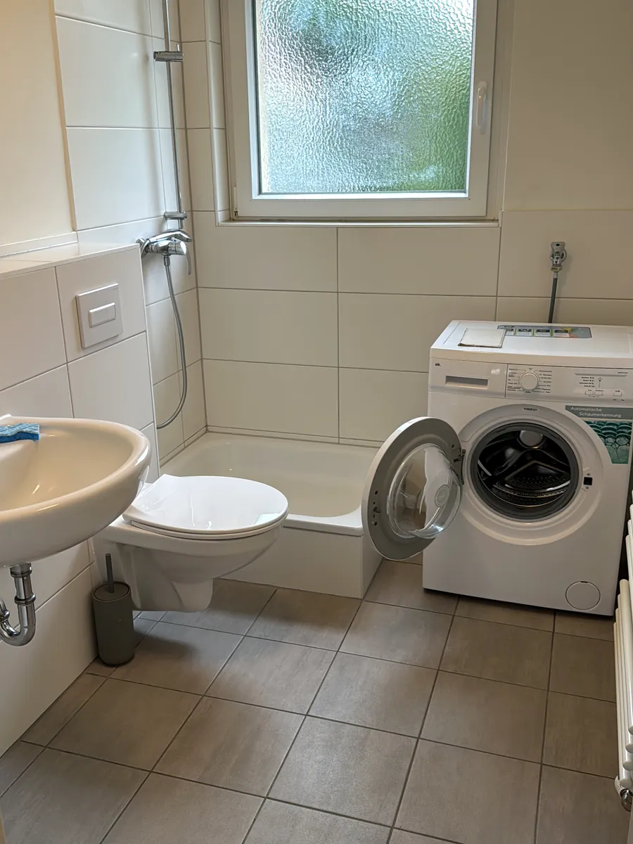 Vorher: kleines Bad mit weißen Wandfliesen, Duschwanne mit Duschstange, WC, Waschbecken und Waschmaschine