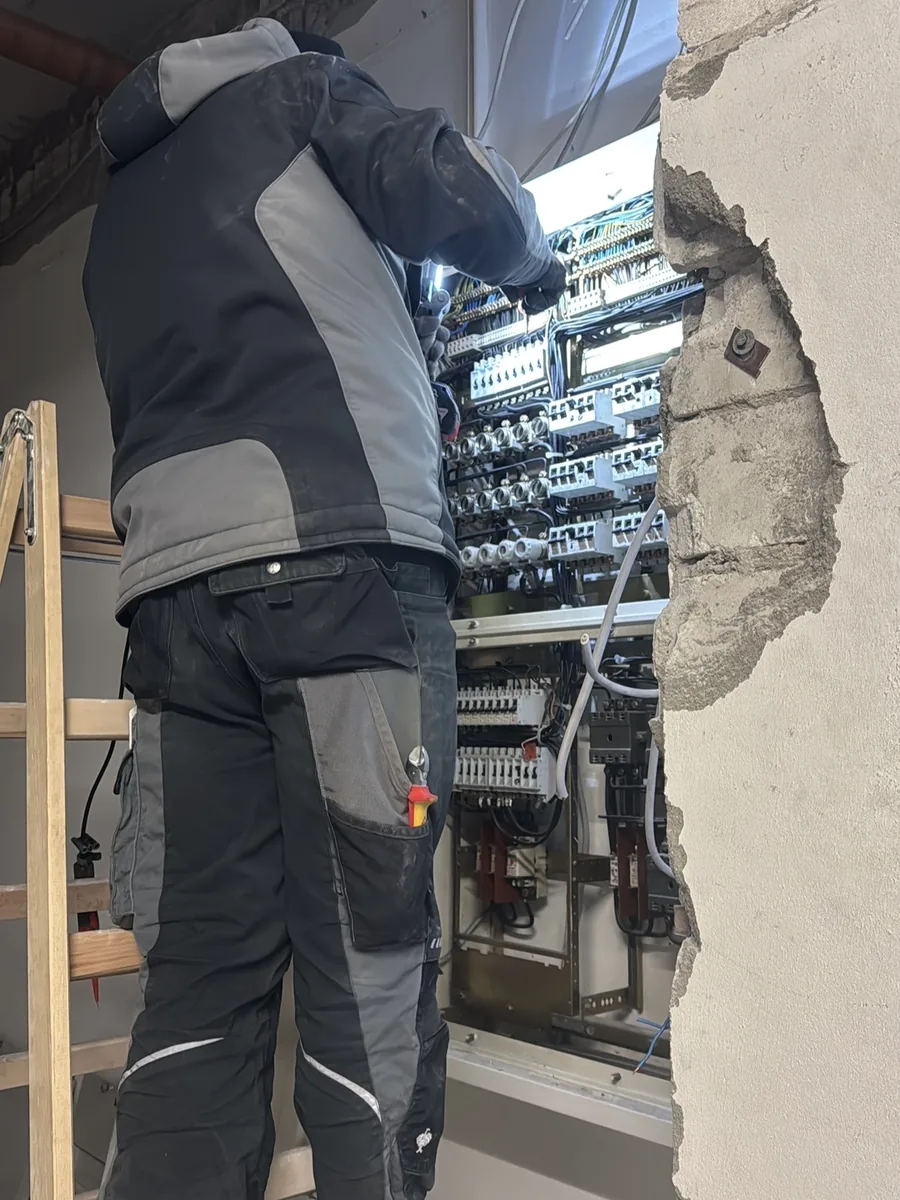 Handwerker auf einer Leiter arbeitet an einem großen Verteiler in einer geöffneten Wand