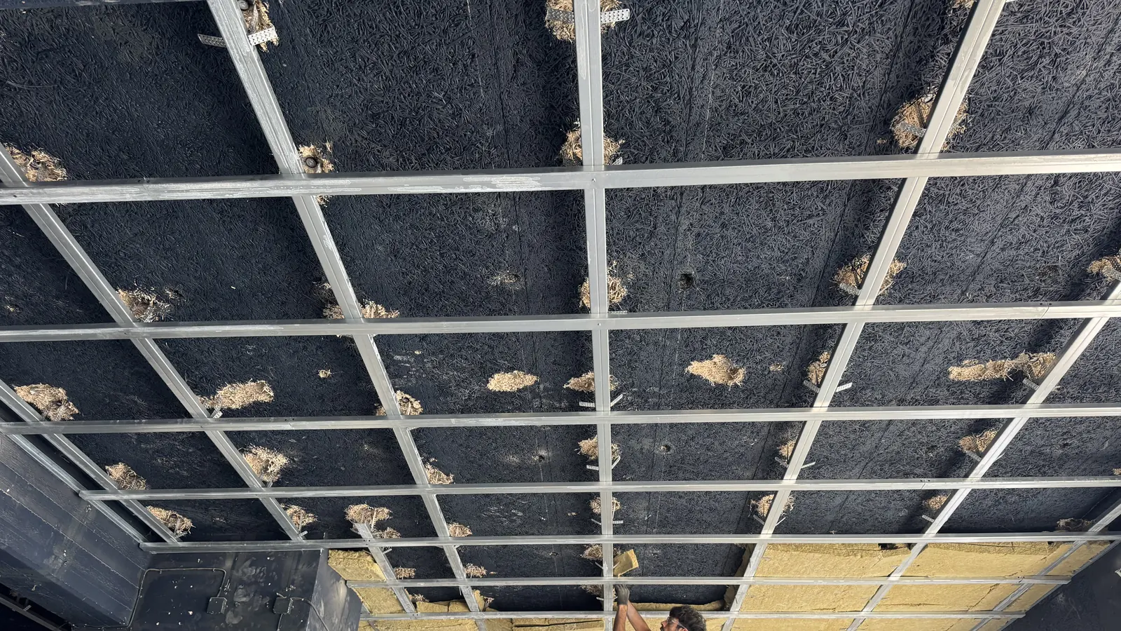 Raster aus Metallprofilen unter einer dunklen Rohdecke, teilweise mit Mineralwolle gedämmt, ein Monteur arbeitet an der Unterkonstruktion