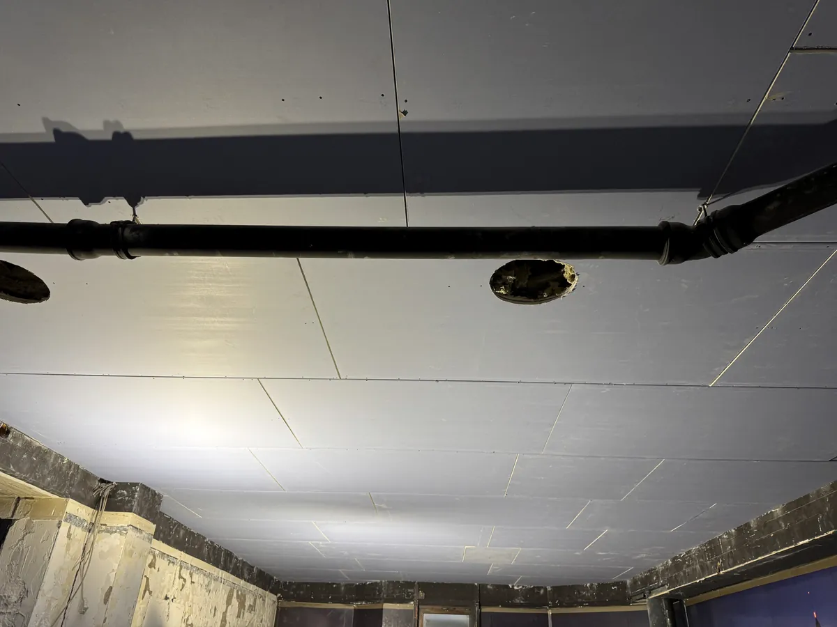 Mit Gipsplatten beplankte Decke vor dem Spachteln, eine Baustellenlampe wirft den Schatten eines Rohrs auf die Decke, die Plattenfugen sind deutlich zu sehen
