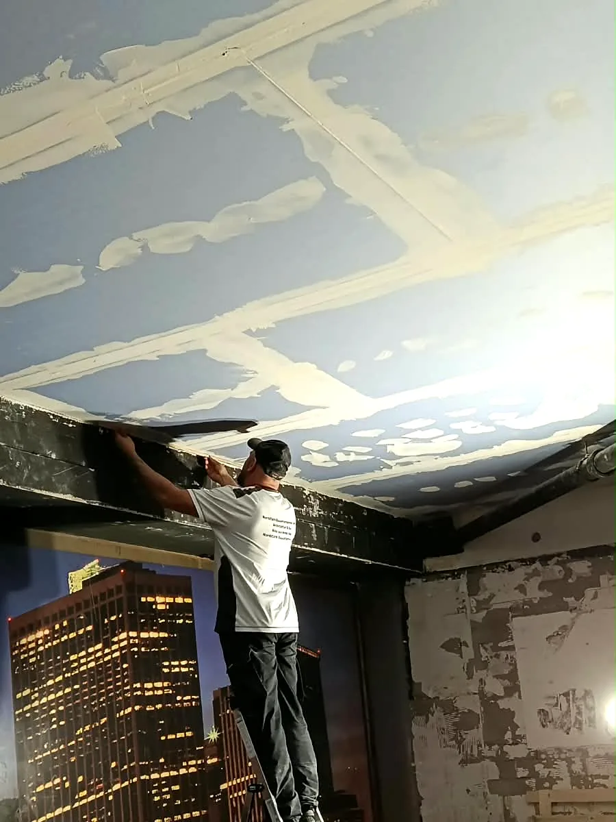 Handwerker auf einer Leiter spachtelt den Randbereich einer neuen Gipsplattendecke, die Fugen sind bereits weiß verspachtelt, darunter eine Wand mit Skyline-Motiv