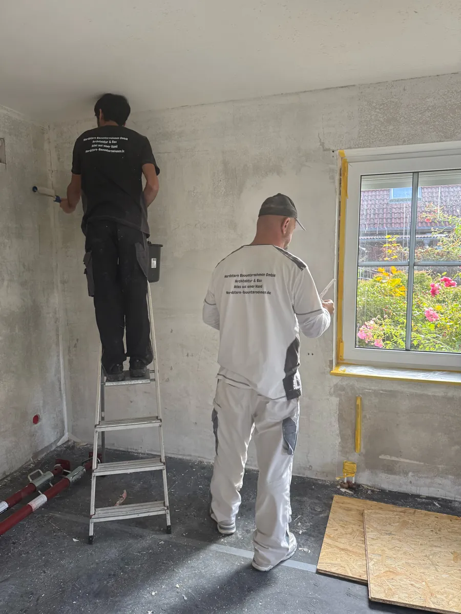 Zwei Handwerker in einem Raum mit Betonwänden: einer steht auf einer Leiter und trägt mit der Rolle Tiefgrund auf, der andere arbeitet am Fenster