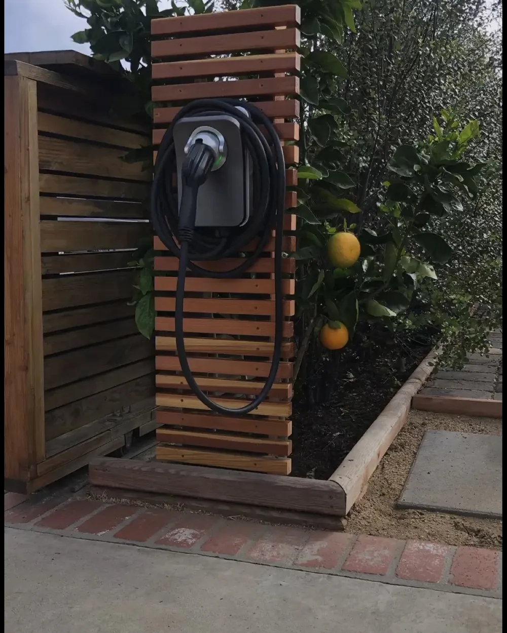 Wallbox mit aufgehängtem Ladekabel an einem Holzlatten-Pfosten neben einer Einfahrt, davor ein Beet mit Zitrusbaum