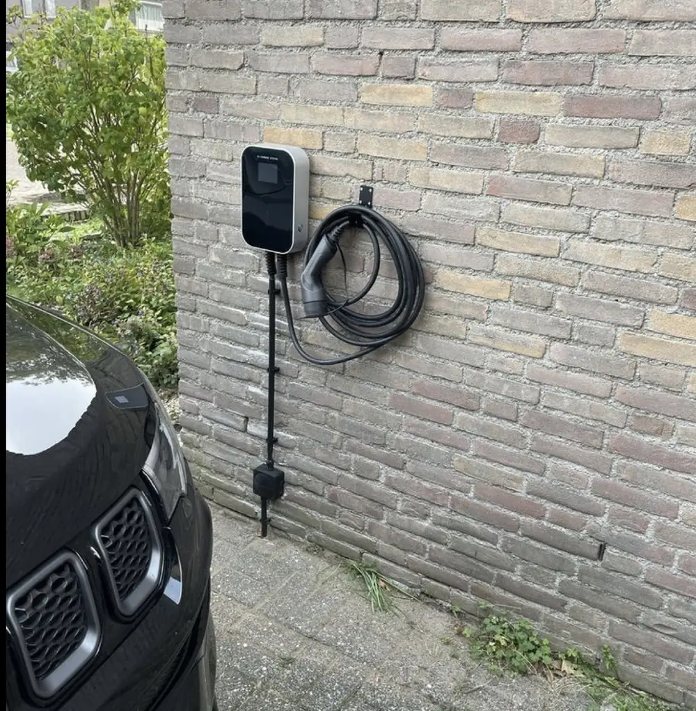Schwarze Wallbox an einer Klinkerwand mit aufgehängtem Ladekabel, Zuleitung in einem schwarzen Rohr nach unten geführt